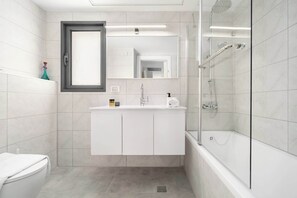 A modern bathroom with a toilet, a sink with a mirror, and a shower area. - Modern 4BR Balcony Mamad Renovated Bldg (Bat Yam)