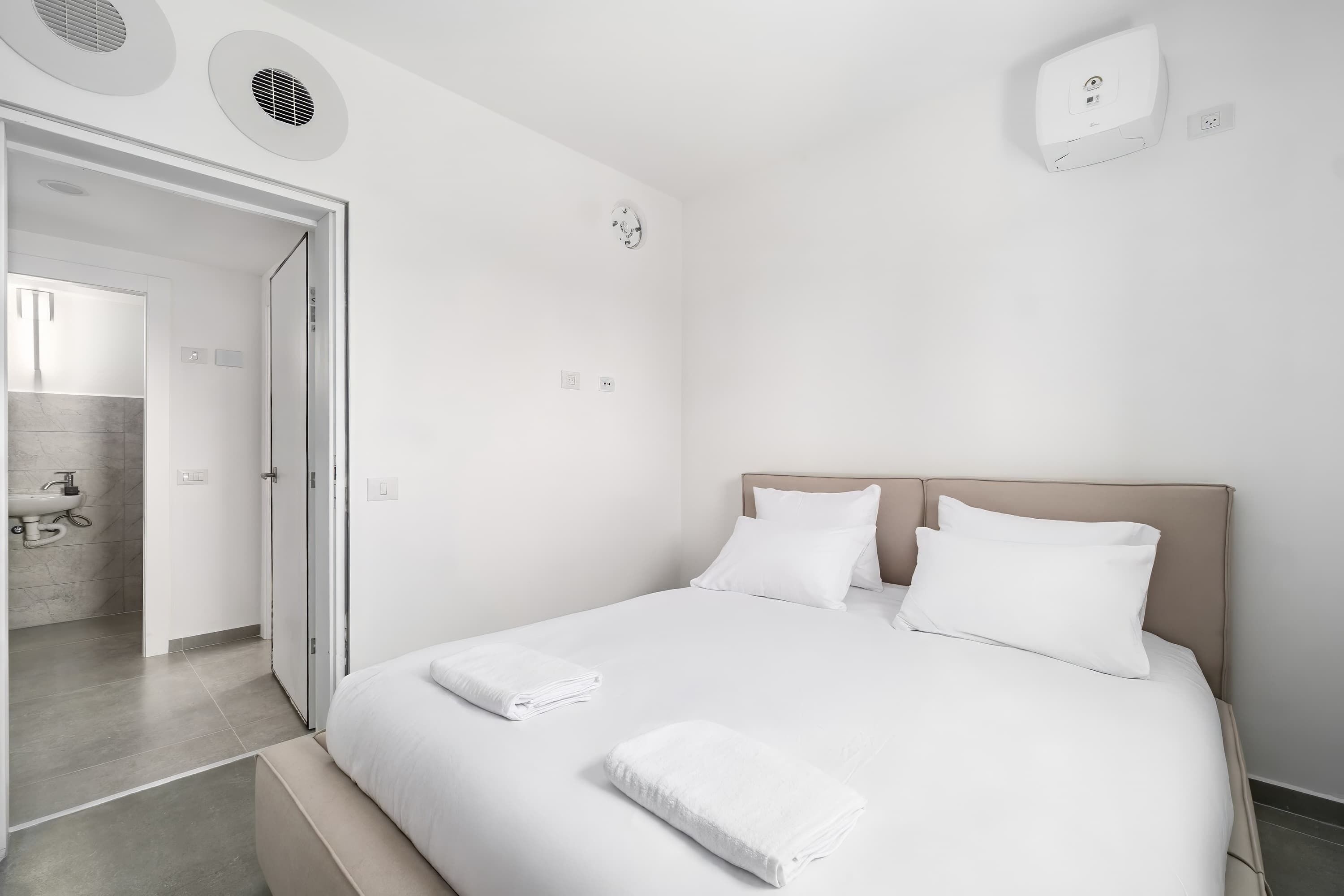 A hotel room with a bed, a bedside table, a wall-mounted alarm clock, and a bathroom visible through an open door.