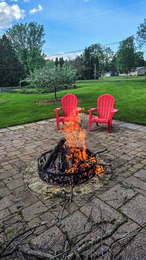 A fire pit with burning logs, surrounded by red chairs, set on a paved patio with a grassy lawn and trees in the background. - Bell Inn Cottage (Sanford)