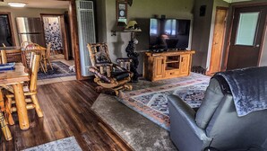 A living room with a wooden floor, a sofa, a TV, a ceiling fan, and a dining area with chairs and a table. - Bell Inn Cottage (Sanford)