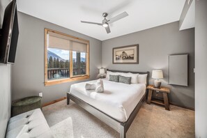 Ein Schlafzimmer mit Bett, Deckenventilator, Fenster mit Blick, Nachttisch mit Lampe und gerahmtem Bild an der Wand.