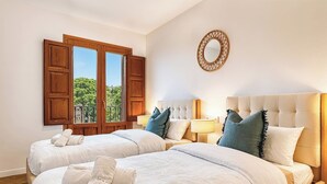 Una camera d'albergo con due letti, un'ampia finestra con persiane di legno, uno specchio rotondo e vista sul verde.