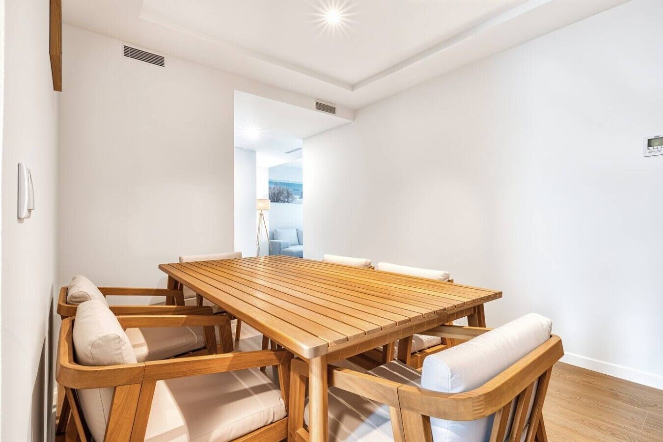 Un comedor con una mesa de madera y sillas acolchadas, un respiradero visible y vista a otra habitación.