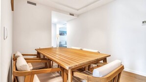 Un coin repas avec une table en bois et des chaises rembourrées, une bouche d’aération visible et une vue sur une autre pièce.