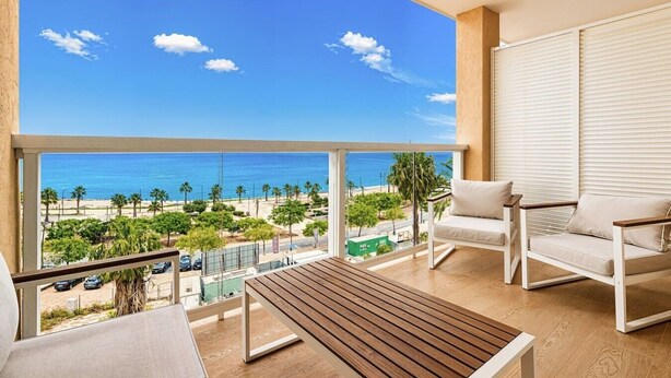 Un balcon avec vue sur la plage, une table en bois et deux chaises blanches.