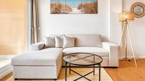 Uma sala de estar moderna com um sofá, uma mesa de centro de vidro, um abajur e uma obra de arte com tema praiano na parede.