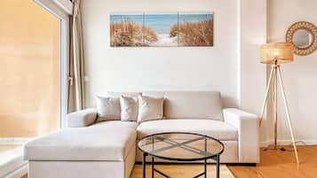 Een moderne woonkamer met een bank, een glazen salontafel, een vloerlamp en een kunstwerk met een strandthema aan de muur.