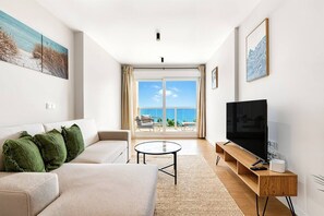 Ein modernes Wohnzimmer mit Sofa, Couchtisch, Fernseher auf Holzfuß und Balkon mit Meerblick.