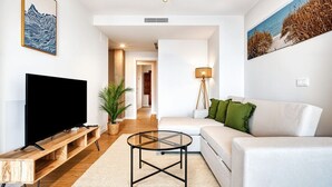 Un salón moderno con un televisor de pantalla plana, un mueble de televisión de madera, una mesa de centro de vidrio, un sofá blanco con cojines verdes y un cuadro de una playa.