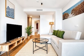 A modern living room with a flat-screen TV, a wooden TV stand, a glass coffee table, a white sofa with green cushions, and a painting of a beach scene. - Sea View Apartment with Terrace - Breeze (La Vila Joiosa)