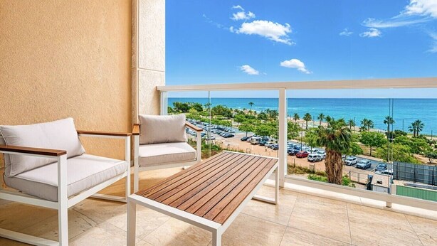 Un balcón con mesa y dos sillas de madera, con vista a una playa con coches aparcados y palmeras.