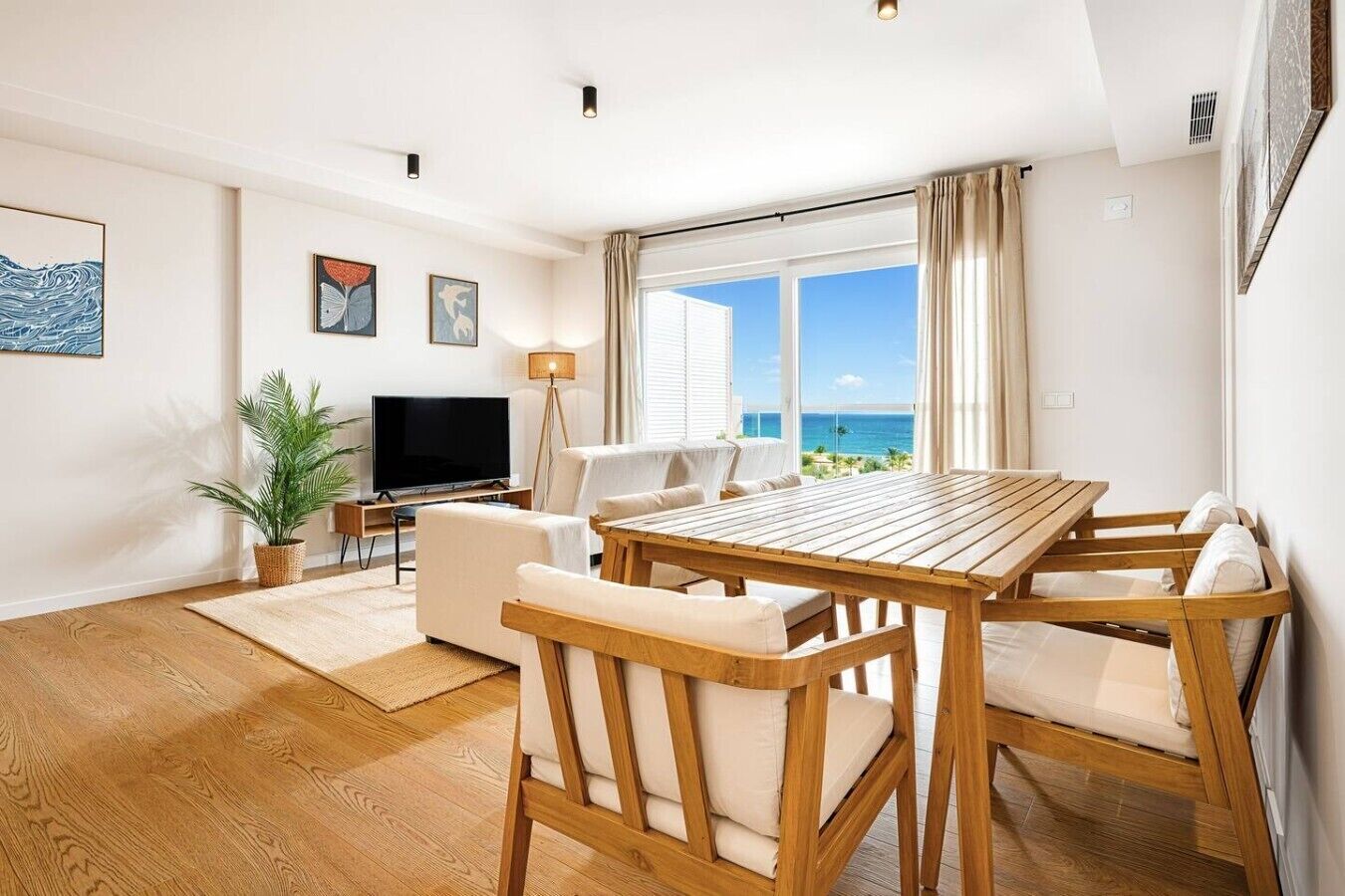 A modern living room with a wooden dining table, white sofas, a flat-screen TV, and a large window with a view of the ocean.
