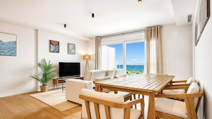 Een moderne woonkamer met een houten eettafel, witte banken, een flatscreen-tv en een groot raam met uitzicht op de oceaan.