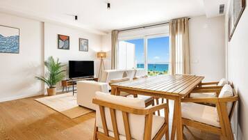 A modern living room with a wooden dining table, white sofas, a flat-screen TV, and a large window with a view of the ocean.