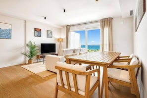 Ein modernes Wohnzimmer mit einem Holzesstisch, weißen Sofas, einem Flachbildfernseher und einem großen Fenster mit Blick auf das Meer.
