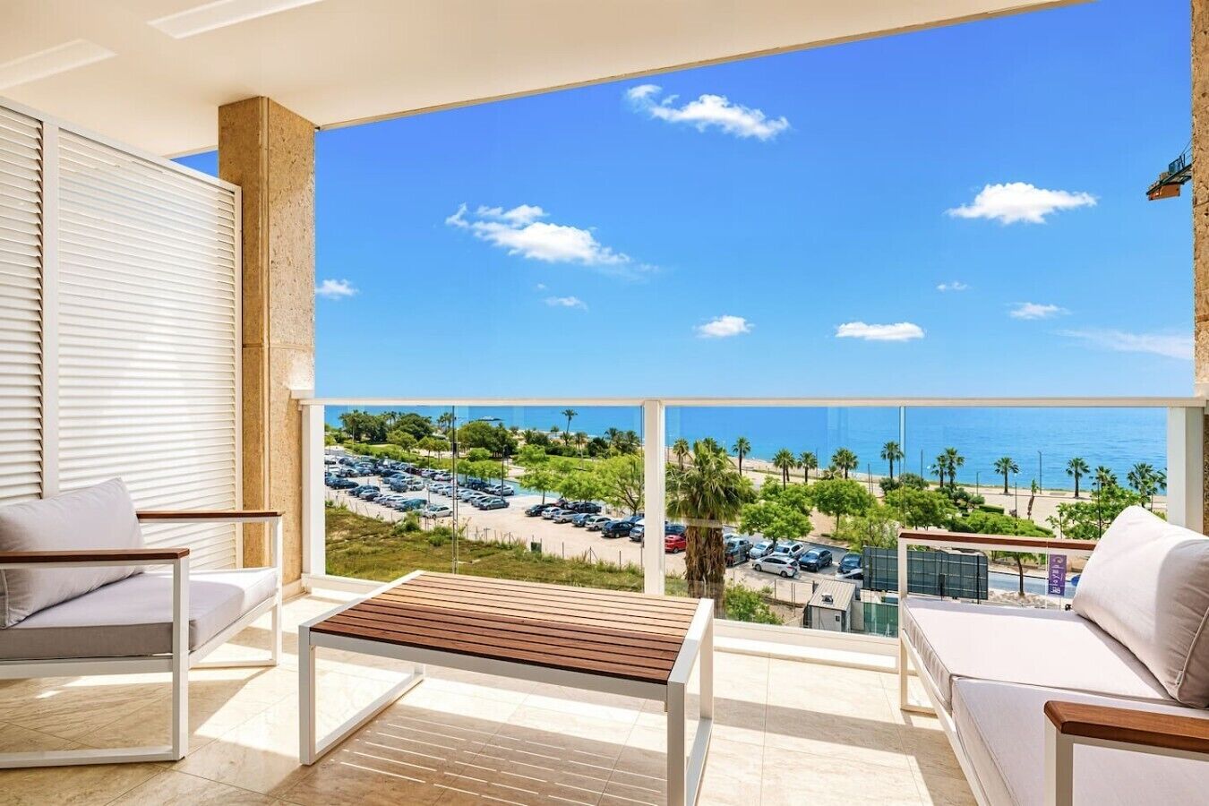 A balcony with a view of the sea, a parking lot, and a building in the background.