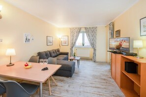 Ein Wohnzimmer mit einer Couch, einem Esstisch mit Früchten und Weingläsern, einem Fernseher und einem Fenster mit Vorhängen.