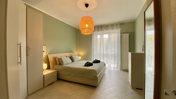 Ein Schlafzimmer mit Bett, Nachttischen, einer Pendelleuchte, einem Fenster mit Vorhängen und einer Tür.