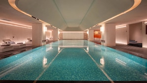 En moderne indendørs swimmingpool med krystalklart vand, omgivet af loungeområder og et geometrisk loftsdesign.