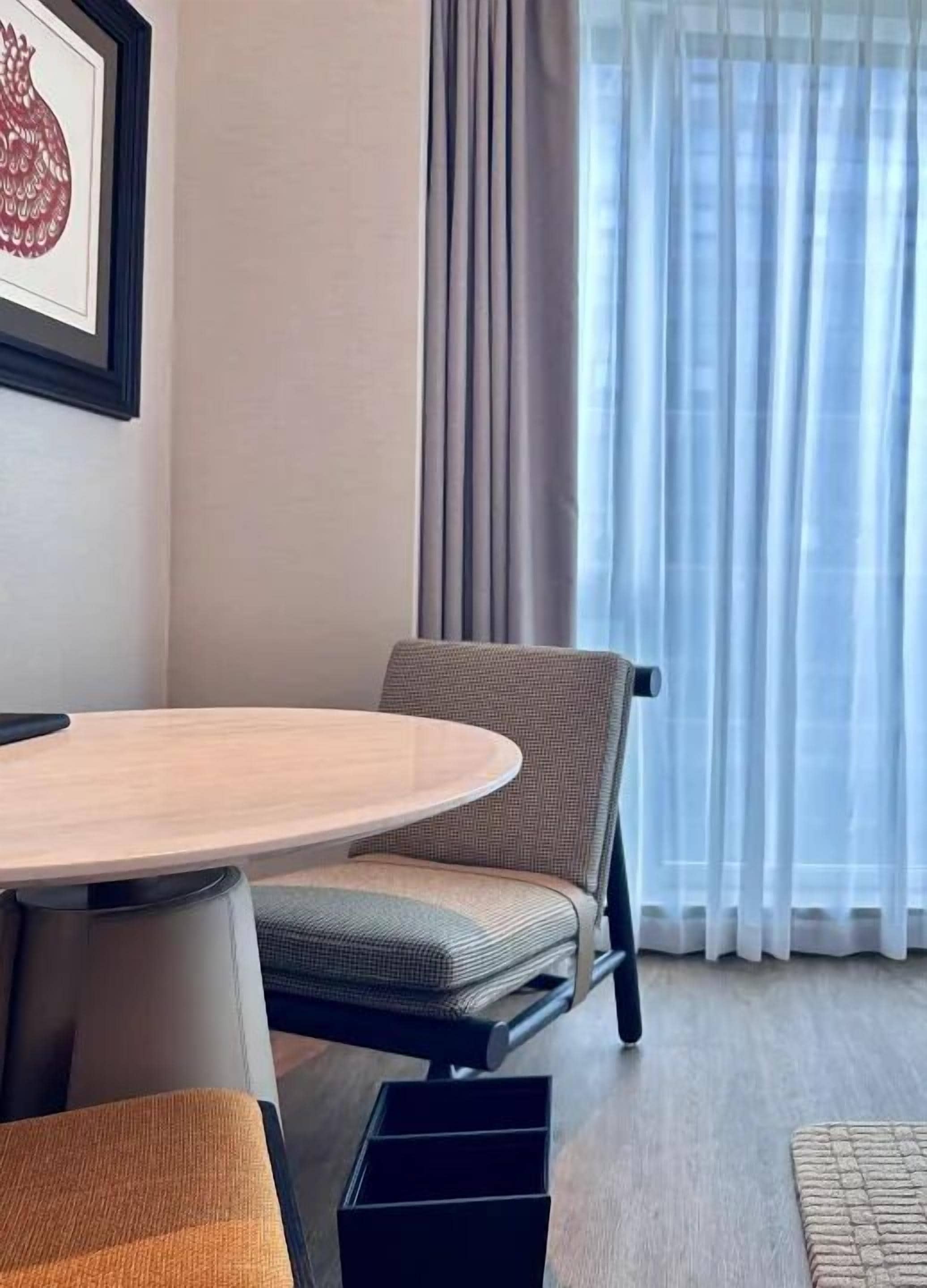 A room with a round table, a chair, a sofa, a framed picture on the wall, and a window with curtains.