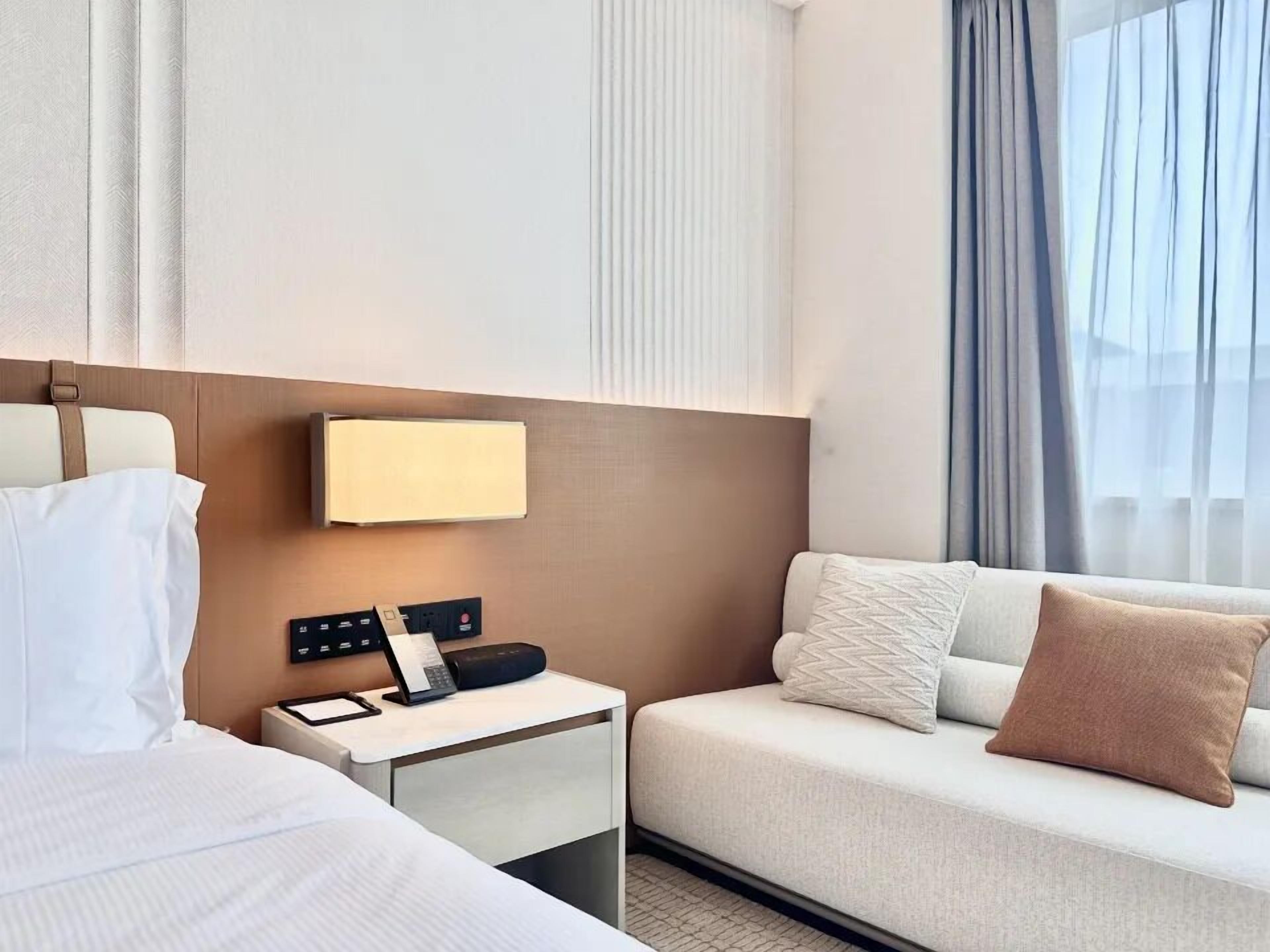 A hotel room with a bed, a bedside table with a lamp, a small desk with a telephone, and a sofa with pillows.
