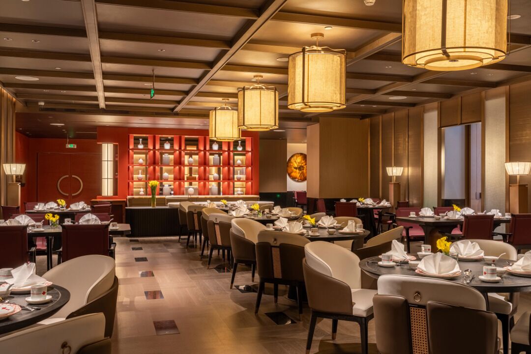 A restaurant with multiple tables and chairs, a red wall with shelves, and hanging lamps.