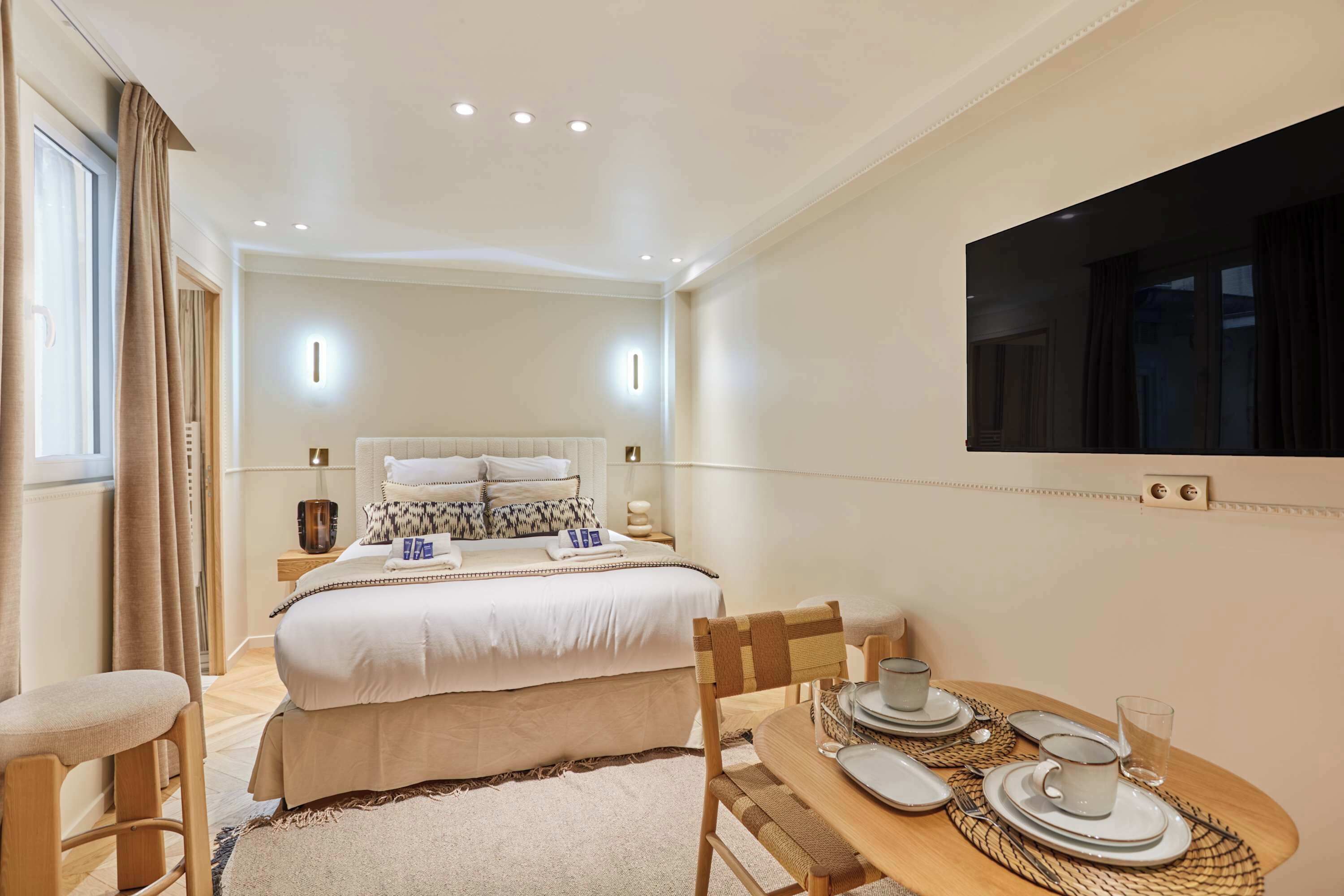 Una habitación de hotel con cama, mesita de noche, lámpara y una mesa de comedor con platos y tazas.