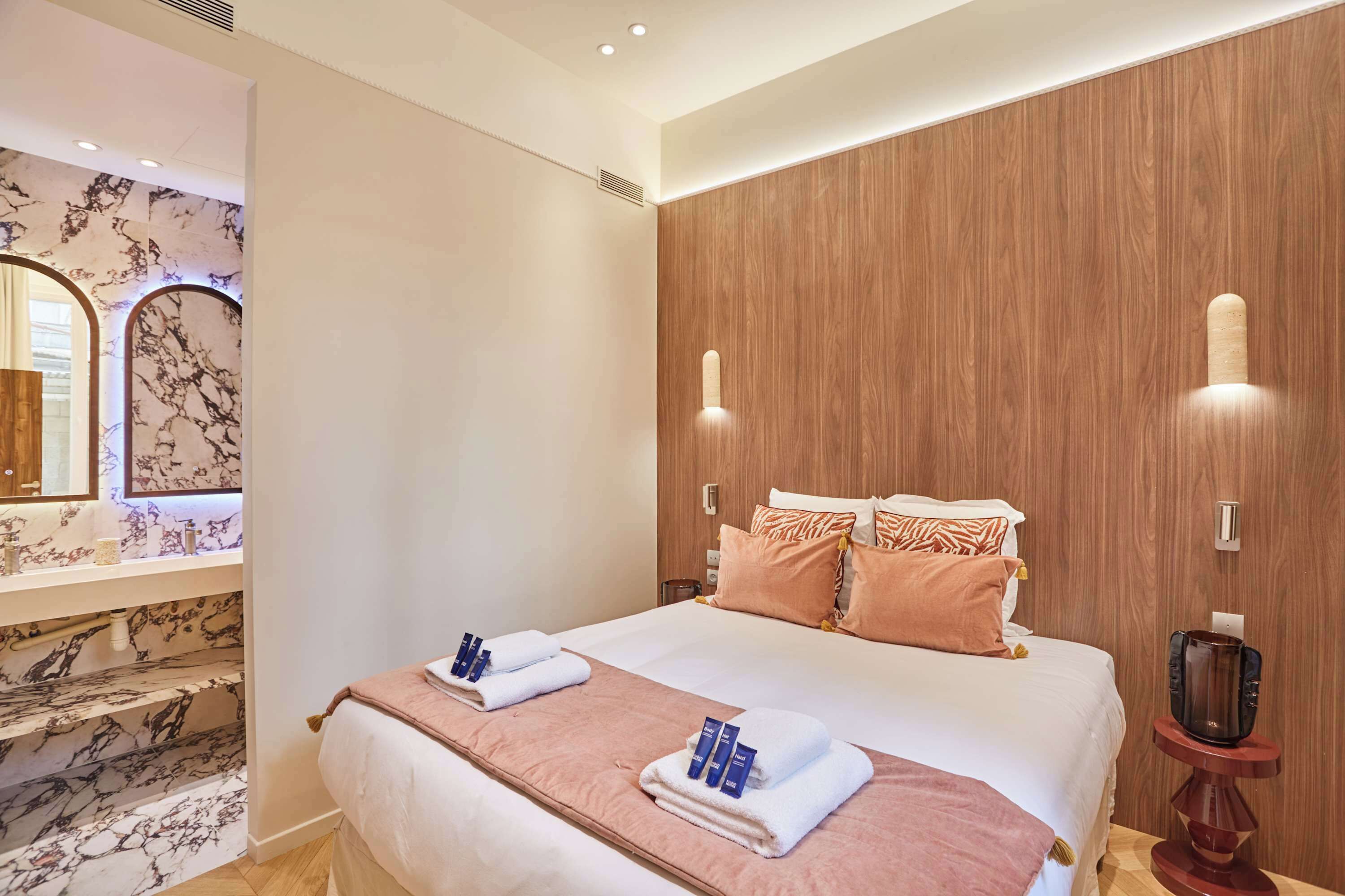 Ein Hotelzimmer mit Marmorbadezimmer, einem Holz-Kopfteil, einem Bett mit Handtüchern und einem Nachttisch mit einer Kaffeemaschine.