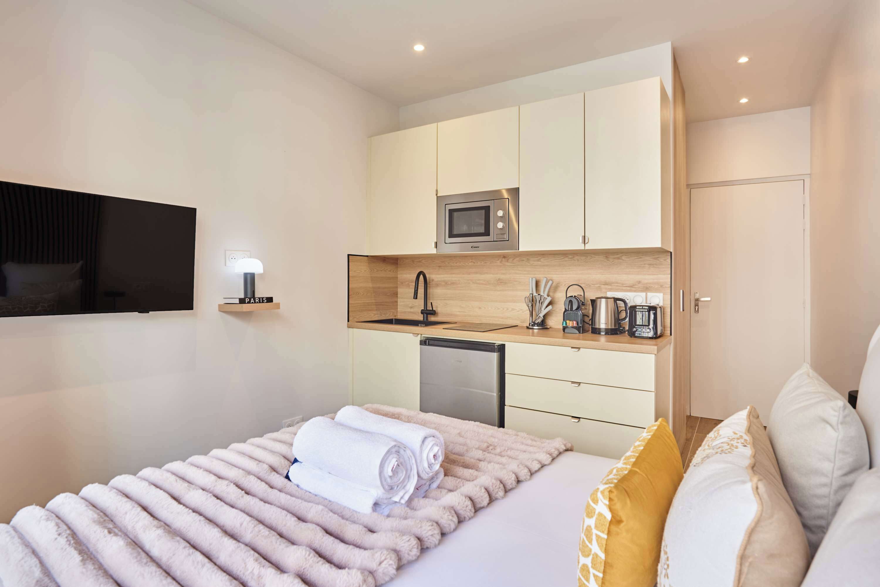 A modern hotel room with a bed, a flat-screen TV, a kitchenette with a microwave, and a small dishwasher.