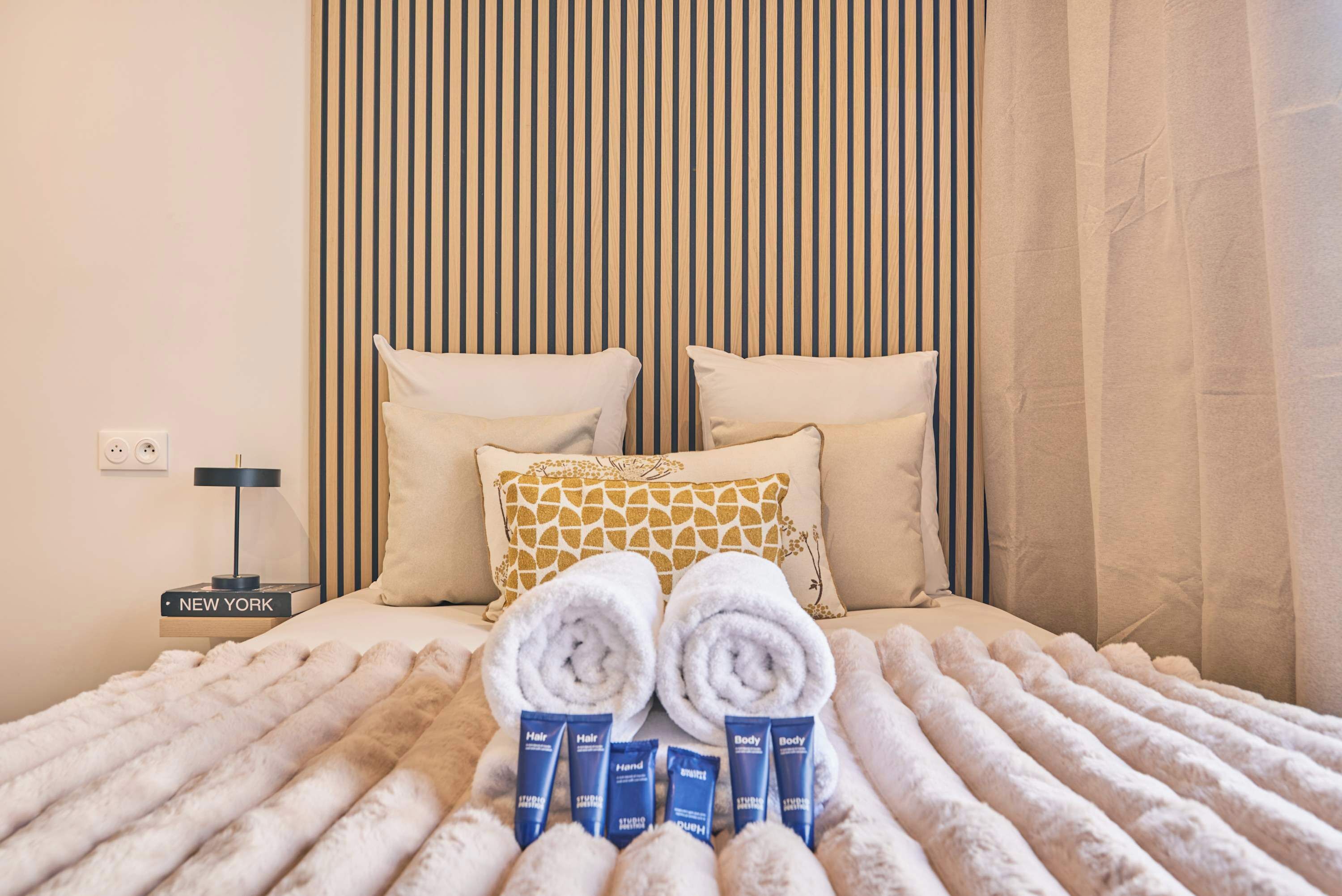 A neatly made bed with rolled towels and toiletries, a bedside table with a lamp, and a striped wall.