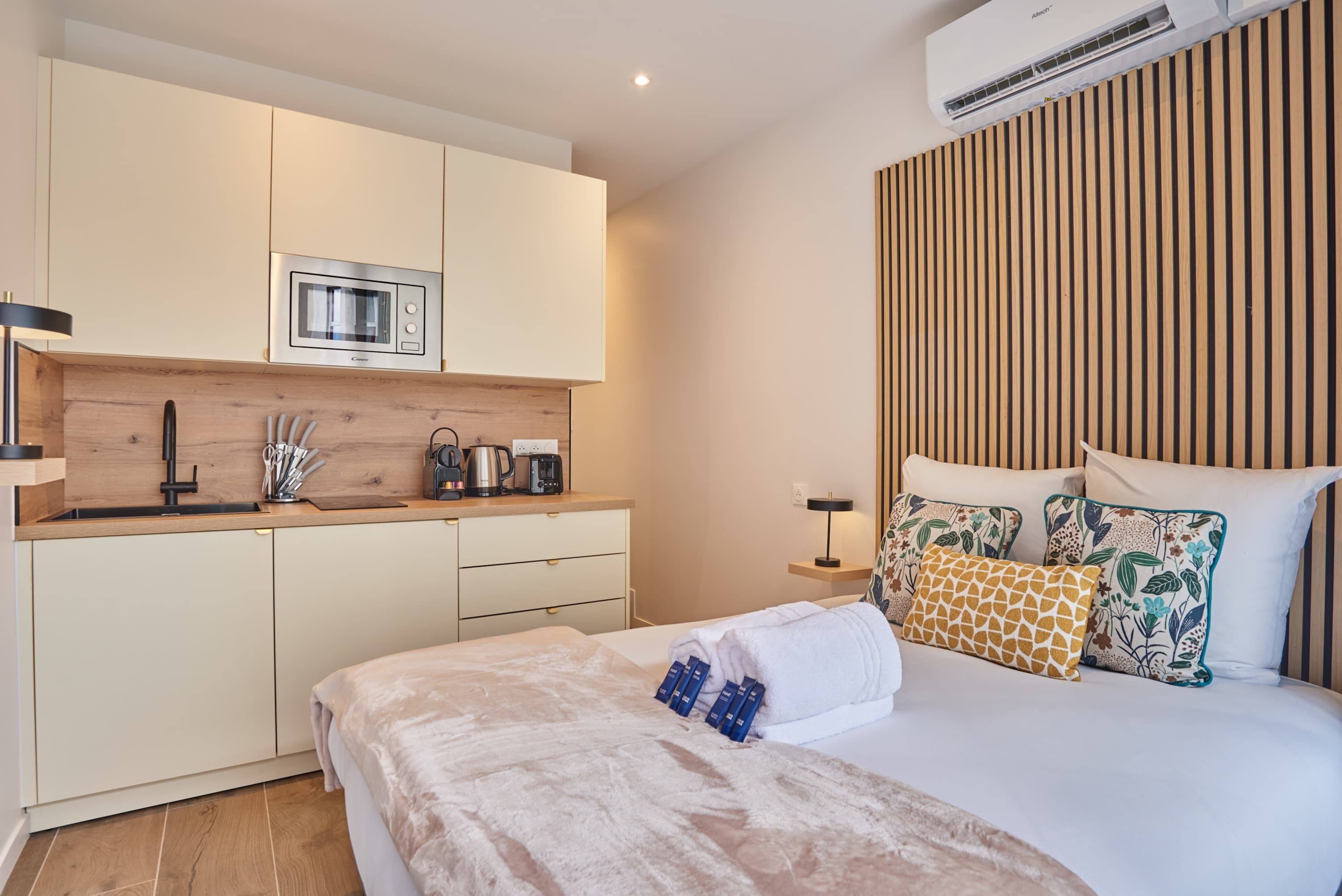 A compact hotel room with a kitchenette, a bed with decorative pillows, and a bedside table with a lamp.
