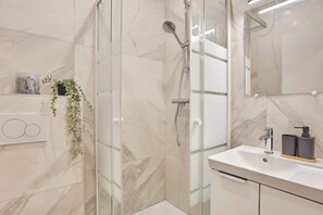 Ein modernes Badezimmer mit einer gläsernen Duschkabine, einem weißen Waschbecken mit schwarzem Seifenspender und einer Wand mit Marmorfliesen.