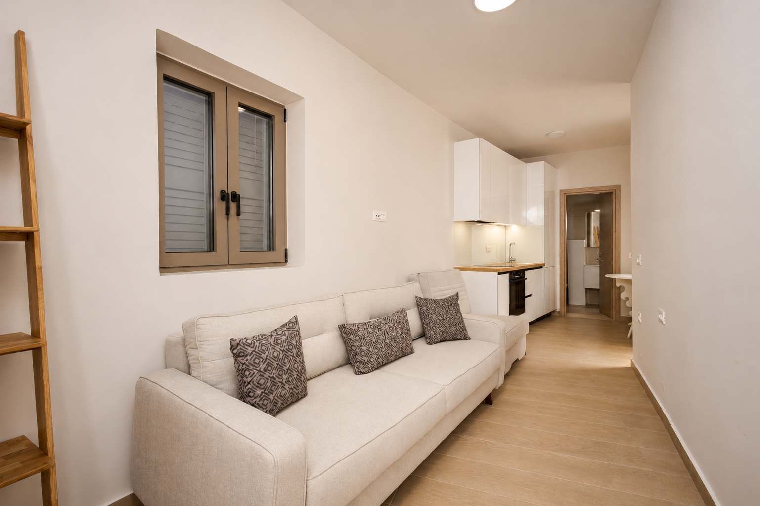 Un appartement moderne comprenant un canapé, des coussins à motifs, une échelle en bois, une cuisine avec des meubles blancs et un couloir menant à une salle de bain.