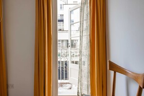 Blick durch ein Fenster mit gelben Vorhängen, einem Holzsessel und einem Gebäude im Hintergrund.