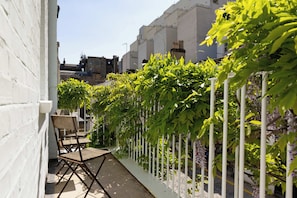 Un étroit couloir extérieur, bordé d’une clôture blanche, avec une chaise pliante et une rangée de plantes vertes.