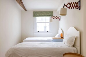 Ein Schlafzimmer mit zwei Betten, einem Fenster mit grüner Vorhangschiene, einer Lampe und einem hölzernen Kopfteil.