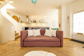 Ein modernes Wohnzimmer mit einem rosafarbenen Sofa, Holzböden, einer Küche im Hintergrund und einer Wendeltreppe.