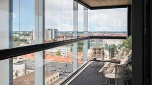 Habitación en un edificio alto con vistas a la ciudad, un balcón con mobiliario para sentarse y un ventanal grande.