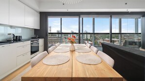 Una cocina moderna con armarios blancos, una mesa de comedor de madera con individuales, y vistas a la ciudad a través de amplias ventanas.