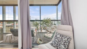 Un salón moderno con sofá, sillón y una mesita, un balcón con vistas a la ciudad y una planta en maceta.