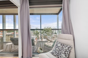 Un salón moderno con sofá, sillón y una mesita, un balcón con vistas a la ciudad y una planta en maceta.