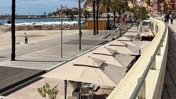 Eine Küstenpromenade mit Palmen, Strandstühlen und Blick auf eine historische Stadt mit Gebäuden und einem Turm.