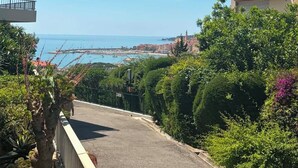 Ein gepflasterter Weg mit Grünpflanzen an beiden Seiten, mit Blick auf eine Küstenstadt und das Meer.