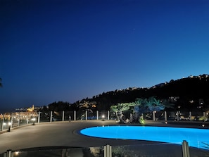 Vue nocturne d’une piscine à bord incurvé, entourée d’une clôture et de palmiers.