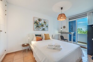 A hotel room with a bed, a small table with a lamp, a painting of palm trees, and a balcony with a view. - LXR Arcadia 2-Bedroom Apartment with Ocean Views (Benalmádena)