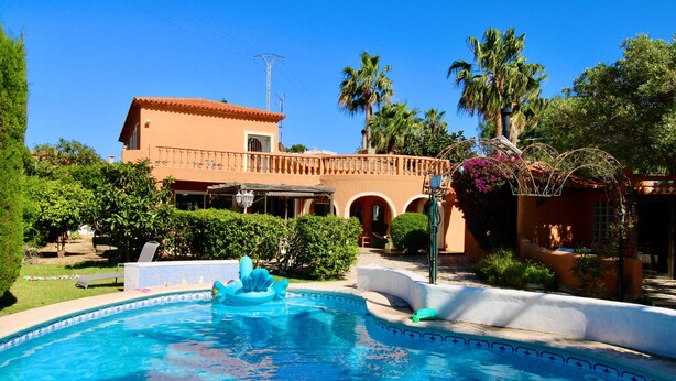 Una piscina con una fuente, rodeada de vegetación y un edificio con techo de tejas.