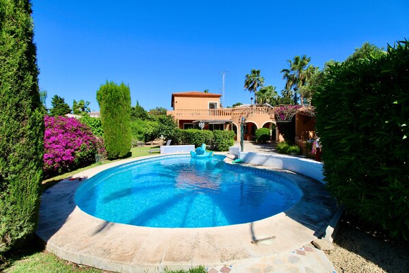 Une piscine avec une fontaine, entourée de verdure, et un bùtiment aux arches en arriÚre-plan.