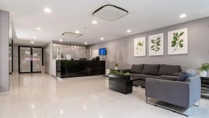 A modern reception area with a glass door entrance, a reception desk, a flat-screen TV, and a sofa with a coffee table. - LKN Grand Ratchathewi (Bangkok)