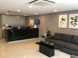 Un área de recepción con mostrador de mármol negro, un sofá gris, una mesa de centro con plantas e ilustraciones botánicas enmarcadas en la pared.