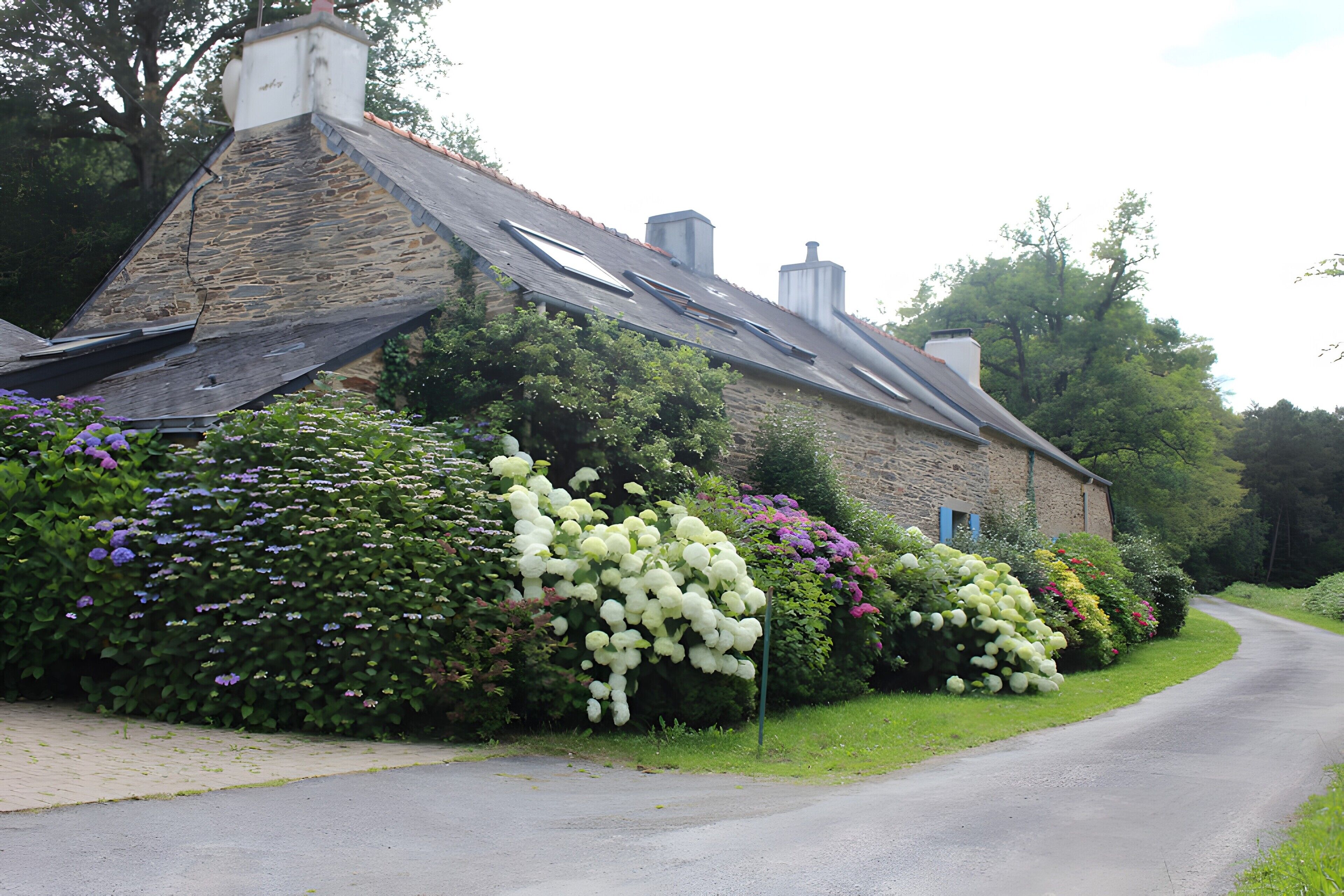 A stone building with a sloped roof, surrounded by lush greenery and flowering plants.
